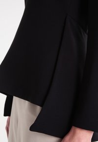 Top nero su misura con una texture liscia, design peplum e orlo curvo. È abbinato a pantaloni di colore chiaro.