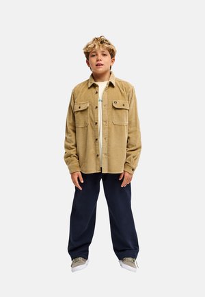 Blond jongetje draagt een zandkleurig corduroy overhemd over een wit T-shirt, donkerblauwe broek en beige sneakers, staand tegen een effen witte achtergrond.