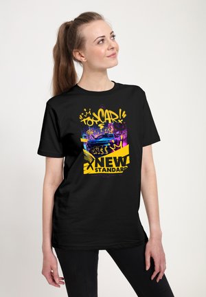 Ung kvinde iført sort t-shirt med farverigt graffiti-stil bil- og street art-design med teksten "NEW STANDARD" i gul og lilla.