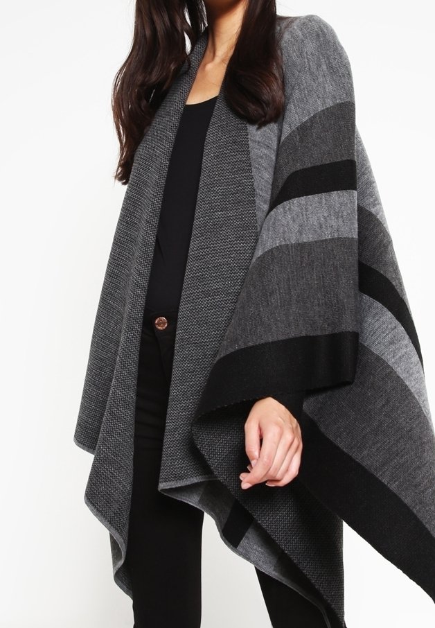 Femme portant un poncho en maille rayé gris et noir par-dessus un haut noir et un pantalon noir, avec de longs cheveux noirs partiellement visibles.