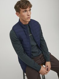 Veste matelassée navy sur un pull vert, avec un col haut et une fermeture éclair. Modèle assis sur un tabouret, portant un pantalon marron.