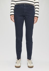 Pantalon en coton slim-fit bleu marine avec cinq poches, fermeture devant par bouton et passants de ceinture, associé à des baskets blanches et beiges.