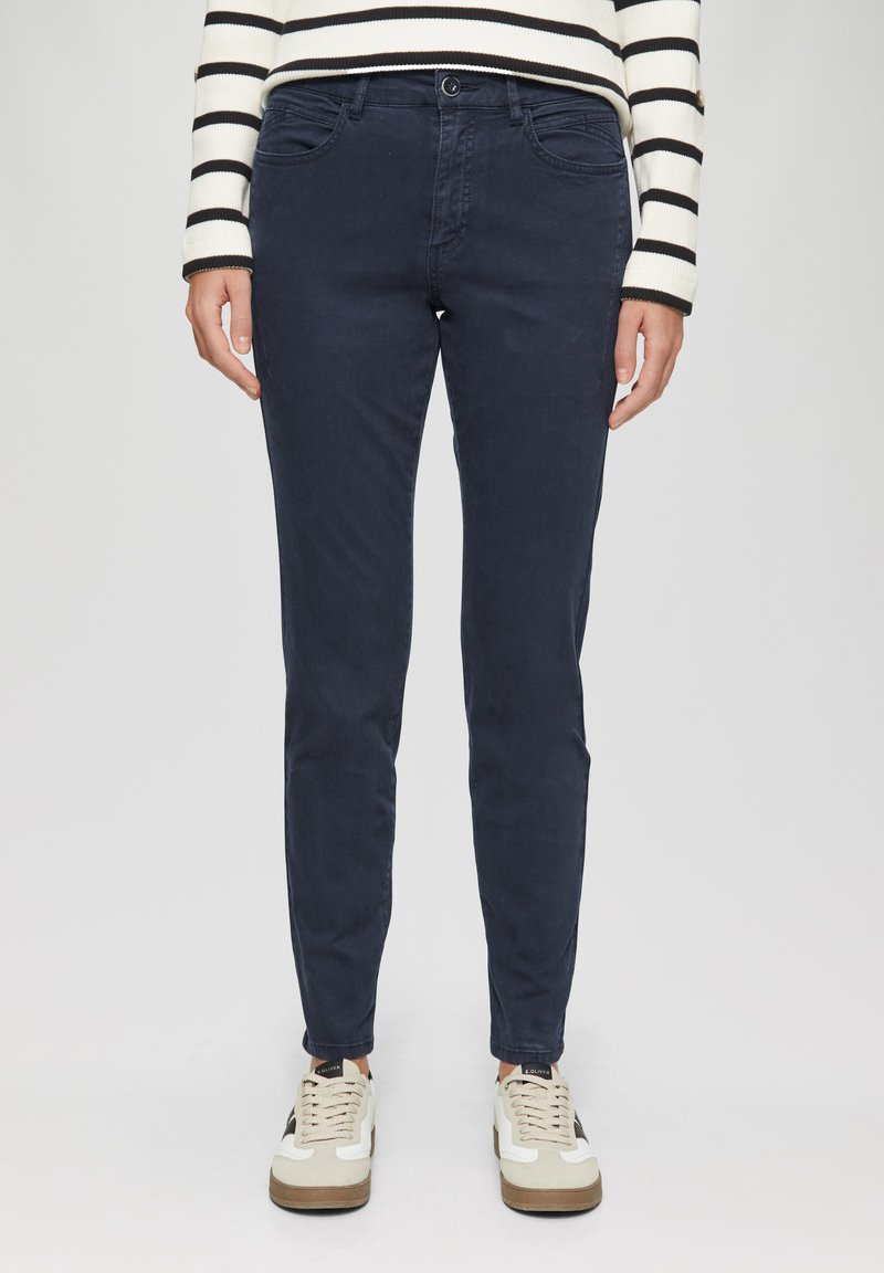 Pantalon en coton slim-fit bleu marine avec cinq poches, fermeture devant par bouton et passants de ceinture, associé à des baskets blanches et beiges.