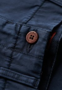 Marineblauw stof met zichtbare textuur, voorzien van een bruine knop met reliëfmerk en een klein zakdetail.