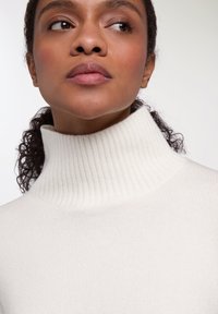 Maglione bianco a collo alto con coste, caratterizzato da un colletto alto, una texture morbida e una silhoutte aderente, incorniciato da capelli scuri e ricci.