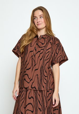 Kobieta o długich blond włosach, ubrana w brązowy zestaw koszuli i spódnicy z abstrakcyjnymi czarnymi falistymi liniami, stojąca na tle jednolitego jasnego tła.