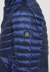 Navy blue puffer jacket with shiny, quilted texture; features a round black logo patch on the left sleeve and a zip closure.