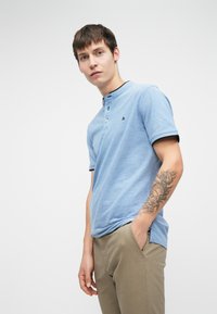 Sininen lyhythihainen polo-paita, jossa on teksturoitu pinta, kahden napin kaulus ja mustat kauluskorostukset, yhdistettynä khaki-housuihin.