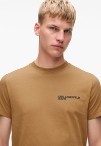 Tričko s krátkým rukávem a kulatým výstřihem v hořčicové barvě z bavlny s hladkou texturou. Na prsou se nachází logo "KARL LAGERFELD JEANS" v tmavém tisku.
