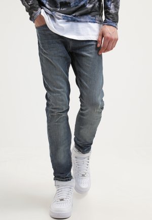 Mężczyzna ubrany w wyblakłe niebieskie dżinsy, białe trampki i wielowarstwową koszulę z górną częścią w kamuflażu, stoi z jedną ręką w kieszeni.