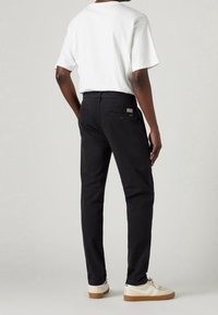 Pantaloni neri in cotone con vestibilità dritta, caratterizzati da due tasche posteriori e un'etichetta con logo. Abbinati a una maglietta bianca e sneakers color crema.