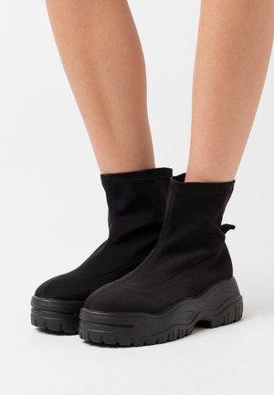 Bottines noires en nylon avec une semelle en caoutchouc épaisse. Elles possèdent un dessus doux et un minimum de coutures, avec une languette pour un enfilage facile.