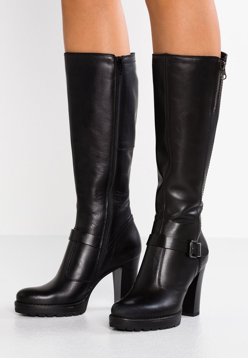 Bottes en cuir noires montantes jusqu'au genou avec talons blocs, fermetures éclair latérales et détail de bride à boucle, portées sur des jambes nues.