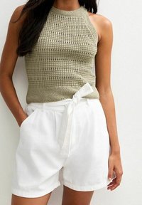 Top de tirantes de ganchillo en verde oliva claro con un cuello alto, combinado con pantalones cortos de lino blancos que tienen un detalle de lazo en la parte delantera y bolsillos laterales.