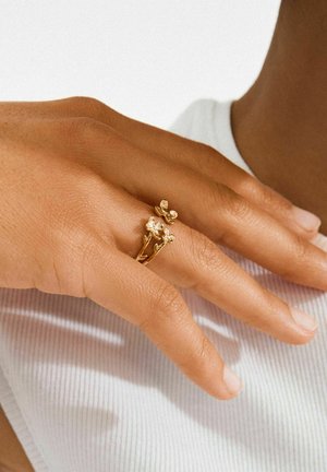 Kuldsetest sõrmustest käel, millel on lillemustrid; nähtav on naha tekstuur. Käsi puhkab valgel kootud riietel, millel on õrn soonikmuster.