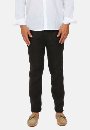 Uomo con pantaloni neri, camicia bianca a maniche lunghe con bottoni e mocassini beige, in piedi con le mani rilassate lungo i fianchi.