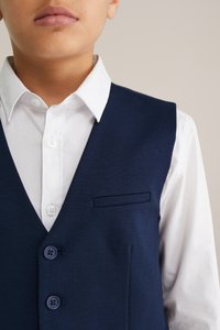 Tengerészkék mellény fehér ingre húzva, V-nyakú kialakítással, két elülső gombbal és egy mellkaszsebbel. Simább szövetes textúra.