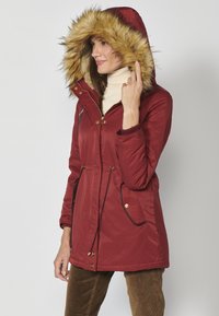 Röd parka-jacka med en huva klädd i konstpeltsfoder, sidofickor, guldbeklädnader och dragsko i midjan. Slät, något glänsande material.