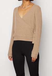 Beige ribbad omslagströja med djup V-hals, långa ärmar och sidoknytning, i kombination med svarta leggings. Egenskapen är mjuk textur.