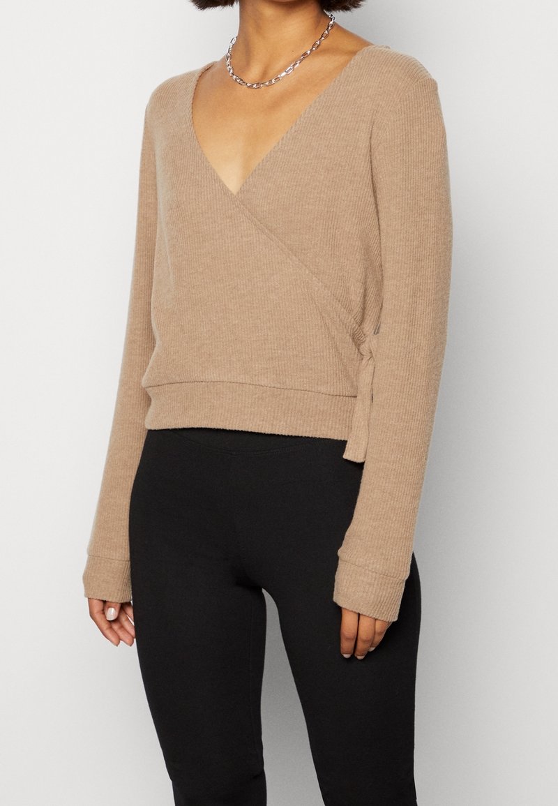 Beige ribbad omslagströja med djup V-hals, långa ärmar och sidoknytning, i kombination med svarta leggings. Egenskapen är mjuk textur.