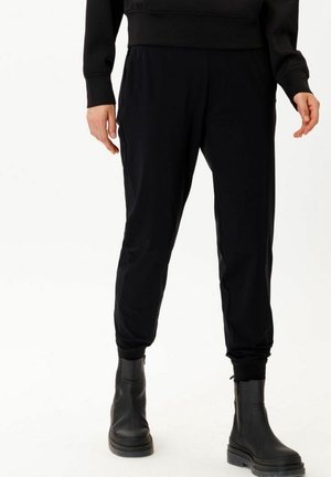Persona con pantalones jogger negros y botas negras al tobillo de pie contra un fondo blanco.