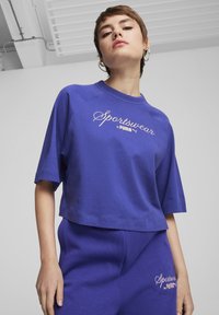 Violet trumpas marškinėliai, pagaminti iš minkštos medvilnės, su apvaliu kaklu ir trumpomis raglan rankovėmis. Balta siuvinėta frazė "Sportswear by PUMA".