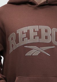 Brūns mikadojums ar lielu, gaiši pelēku "REEBOK" logo un mazu logo akcentu. Izgatavots no mīkstas auduma ar teksturētu apdari un priekšējo kabatu.