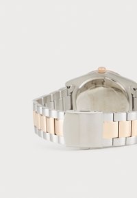 Silberne und roségoldene Metalluhr mit einem Gliederband, das eine polierte Oberfläche aufweist, sowie einemverschluss, der mit dem Markennamen "GUESS" graviert ist.