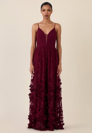 Frau in einem ärmellosen, tief burgunderroten Kleid mit floralen Applikationen und tiefem Ausschnitt, stehend vor einem schlichten beigen Hintergrund.