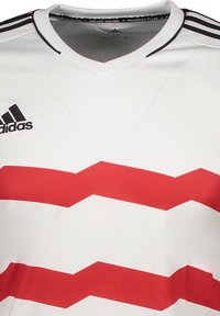 Witte sporttrui met een V-hals, voorzien van rode zigzagstrepen. Zwart Adidas-logo op de linkerborst en twee zwarte strepen op de schouders.