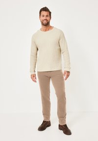 Beige Waffelstrick-Langarmshirt, beige Cordhose und dunkelbraune Schnürstiefel. Stehendes männliches Modell vor neutralem Hintergrund.