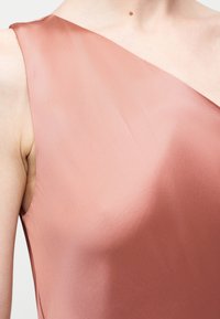 Femme portant un haut en satin sans manches à une épaule de couleur rose poudré, dévoilant le cou, l'épaule et le haut de la poitrine.