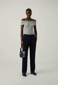 Sieviete ģērbta pelēkā off-shoulder topā, tumši zilos biksēs, melnās norādītajās kurpēs, turamā mazā melnā rokassomiņā pret vienkāršu gaišu fona.