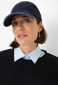 Navy baseball cap with light blue accent, worn with a light blue collared shirt under a black sweater. Features a close-up face view.