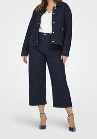 Mörkblå denimjacka med fickor, i kombination med vida mörkblå jeans. Vit V-ringad topp under. Blå högklackade skor.