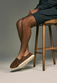 Braune Wildleder-Loafers mit weißen Gummisohlen und Schnürdetails. Die Schuhe werden von einer Person getragen, die auf einem Holzschemel sitzt.