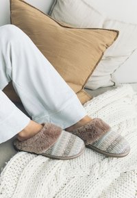 Pantofole lavorate a maglia con motivo multicolore; bordo in pelliccia marrone morbida e suola in gomma. Indossate con pantaloni bianchi larghi su una coperta chiara.