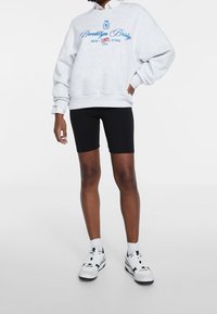 Felpa grigia con testo blu "Brooklyn Bridge", pantaloni corti neri da bicicletta e sneakers bianche, presentati su uno sfondo neutro.