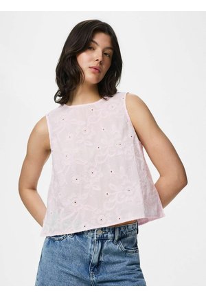 Jeune femme aux cheveux foncés portant un haut sans manches rose clair brodé de fleurs et un jean en denim bleu, posant devant un fond blanc.