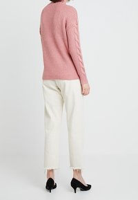 Rosa stickad tröja med kabelstickningsdetaljer på ärmarna, tillsammans med krämfärgade byxor som når anklarna och svarta klackskor.