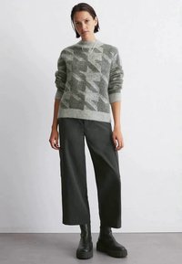 Woman wearing gray patterned sweater, black wide-leg pants, and black chunky boots, standing against a plain white background.