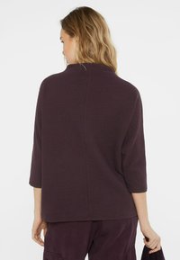 Pflaumenlila Langarmshirt mit hohem Kragen, strukturiertem Stoff und lockerer Passform. Sichtbare Naht am Rücken, kombiniert mit passenden Hosen.