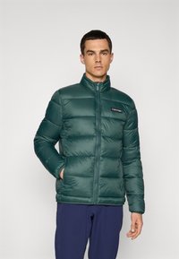 Man wearing a dark green quilted Calvin Klein jacket with zip pockets and navy blue pants, standing against a plain white background.