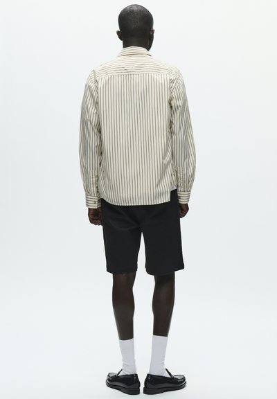 Chemise beige rayée à manches longues avec des lignes noires, short noir, chaussettes blanches et chaussures noires. Vue de dos sur un fond uni.
