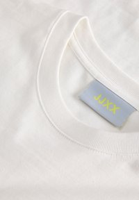 Κοντινή λήψη ενός λευκού T-shirt με στρογγυλή λαιμόκοψη που δείχνει ριμπ λαιμόκοψη και μια γκρι ετικέτα με κίτρινο κείμενο "JJXX".
