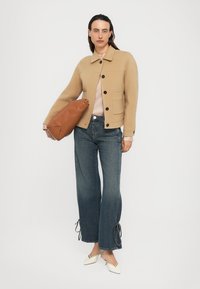 Frau in beiger Knopfjacket, hellrosa Oberteil, weit geschnittenen blauen Jeans mit Bindebändern, weißen Kitten-Heels, hält eine große braune Tasche vor weißem Hintergrund.