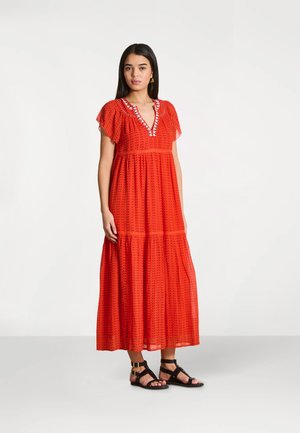 Femme debout portant une robe rouge à motifs longue jusqu'aux chevilles avec des manches volantées et des sandales plates noires à lanières sur un fond uni.
