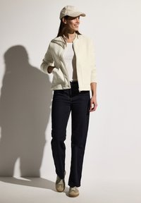 Veste zippée beige clair, chemise blanche, jeans bleu marine et baskets beiges. Les accessoires incluent une casquette beige clair. Design minimaliste.