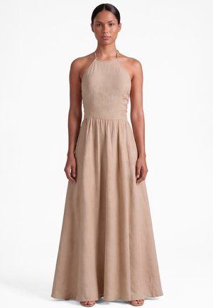 Kobieta stojąca przodem, ubrana w bezrękawnikową beżową sukienkę maxi z halterem, dopasowanym górnym partem i płynną spódnicą.
