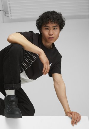 Maglietta grafica in bianco e nero con lettering audace, abbinata a jeans di denim neri e sneaker nere. La persona è accovacciata, con capelli ricci.